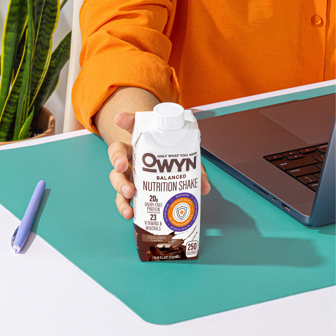Person holding a OWYN Chocolate Balanced Nutrition Shake in front of a laptop on a teal desk.