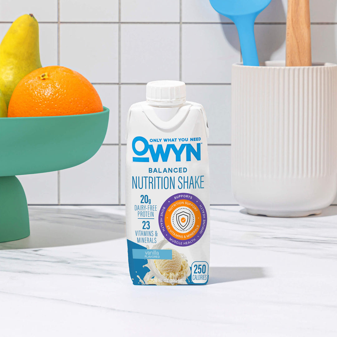OWYN Vanilla Balanced Nutrition Shake package on a kitchen counter with fruits and utensils in the background
