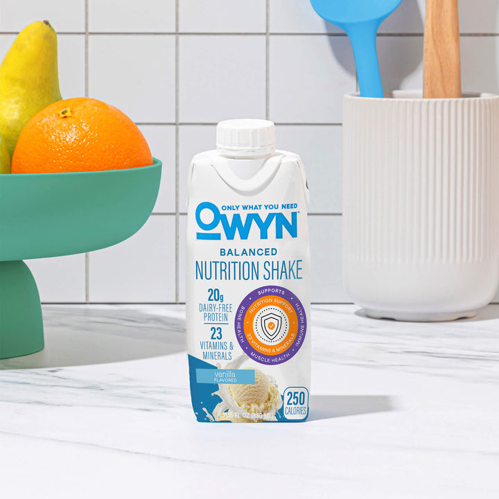 OWYN Vanilla Balanced Nutrition Shake package on a kitchen counter with fruits and utensils in the background